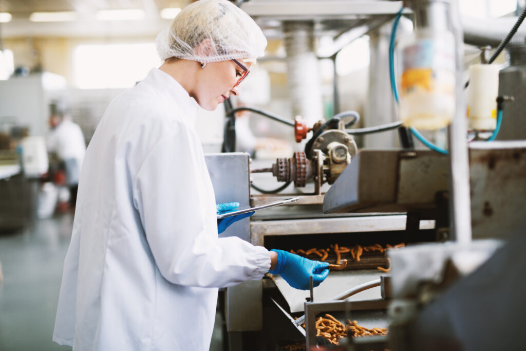 Young female worker in sterile clothes is taking a sample of salt snacks from production line.